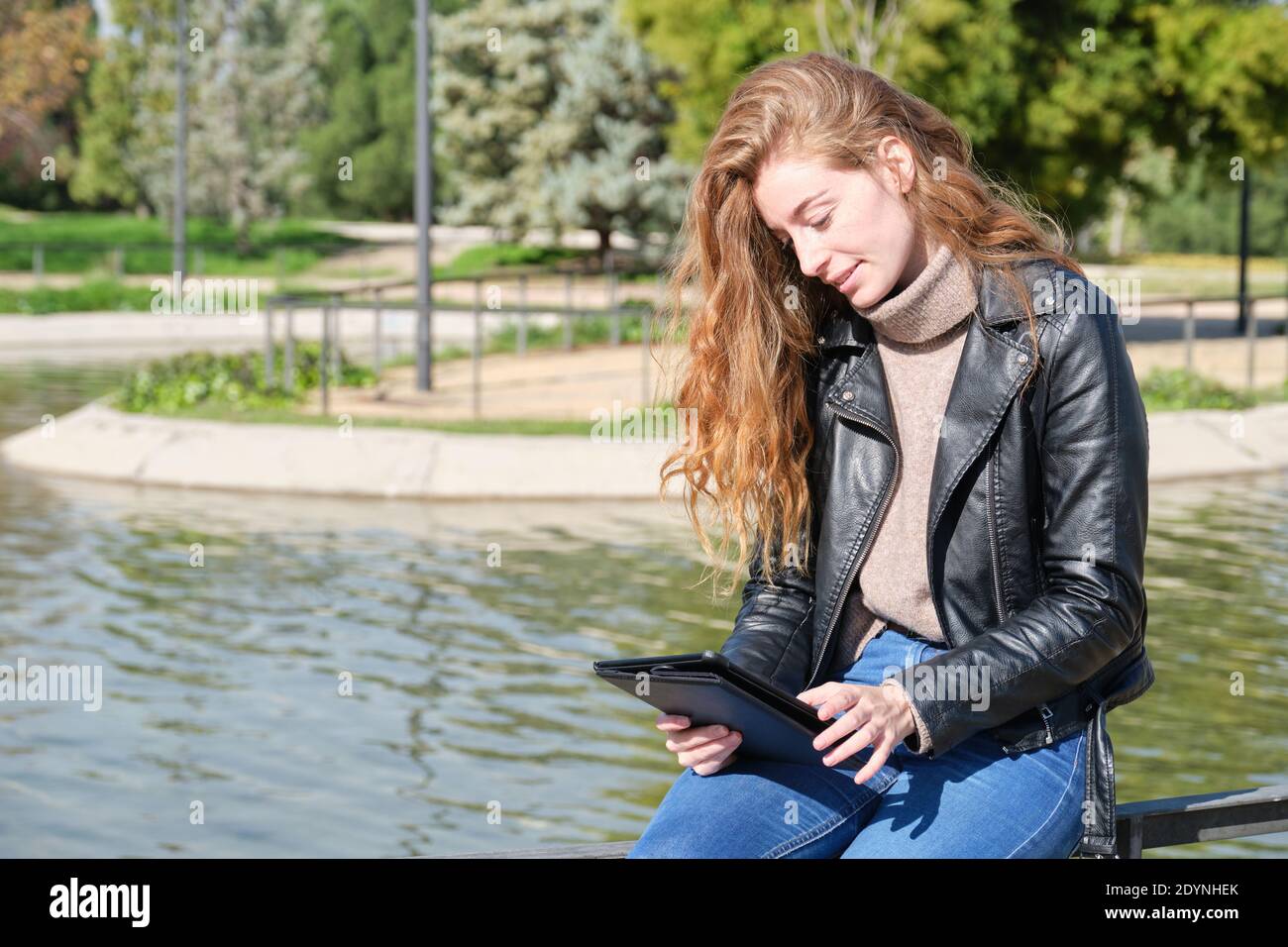 Redhead caucasian woman reading or working in a e-book or tablet in a park. Digital nomad entrepreneur concept. Stock Photo