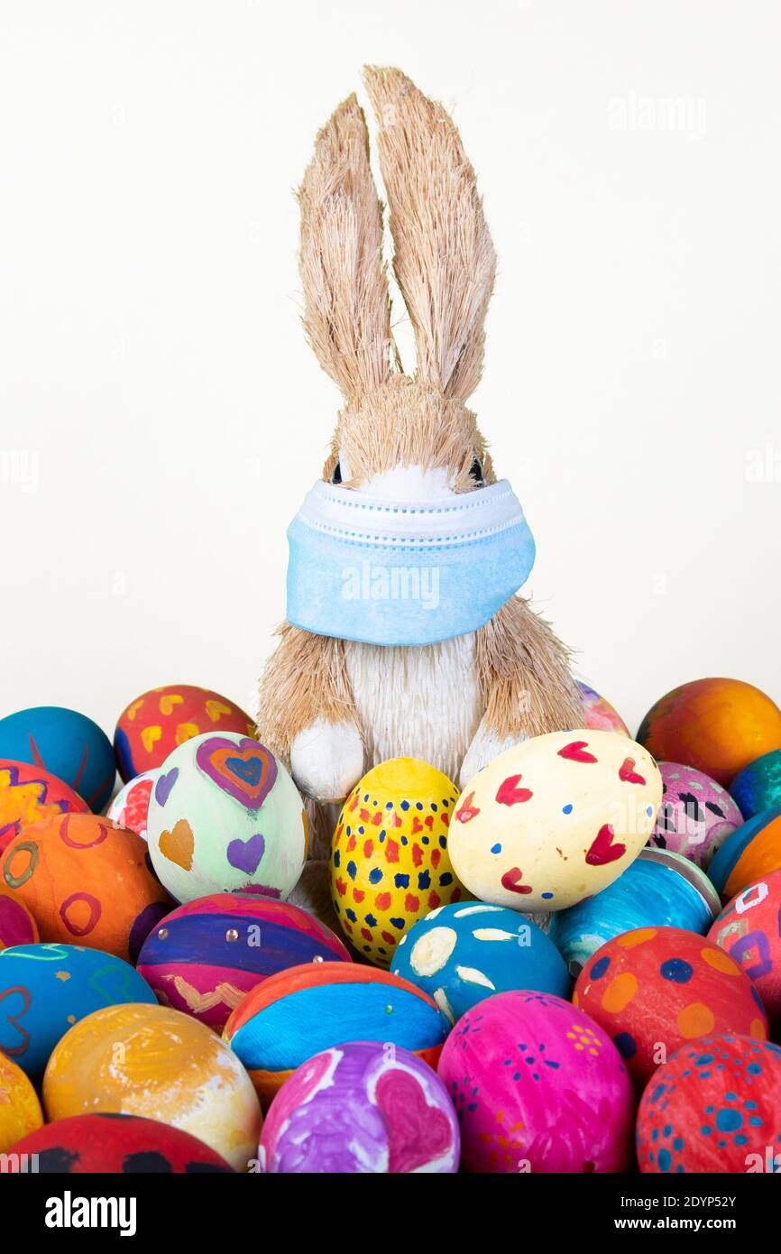 rabbit figurine with face mask between painted easter eggs before white background, symbol for impact of corona virus on easter season Stock Photo