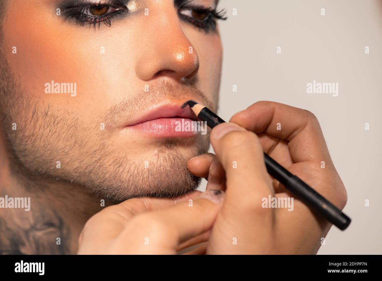Hand of contemporary makeup artist with black kohl eye liner going to apply it on lips of male fashion model sitting in front of camera Stock Photo