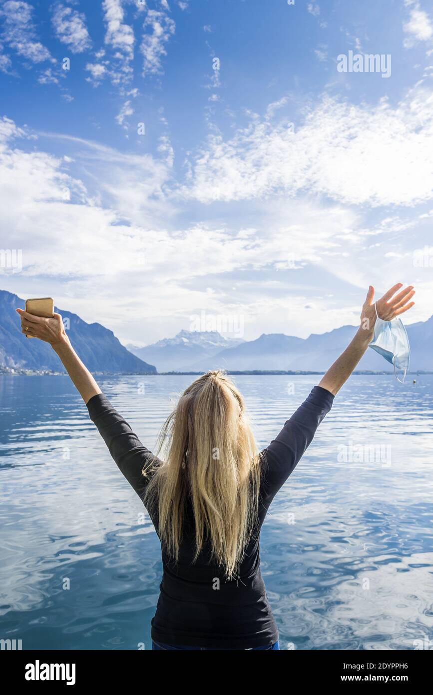Vertical photo of a blonde woman with her arms raised representing freedom with a mobile phone in her hand and a mask in the other hand in front of an Stock Photo