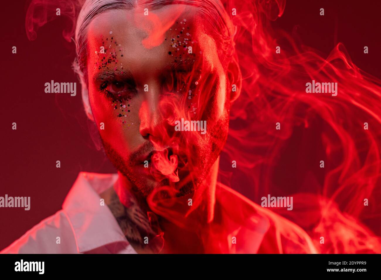 Young man with stage makeup, white hair in accurate hairdo and tattoo on neck looking at you while standing in front of camera and smoking Stock Photo