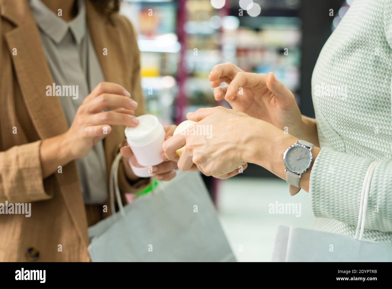 Hands of two young elegant consumers with paperbags applying cosmetic cream while testing it in beauty shop and choosing which to buy Stock Photo