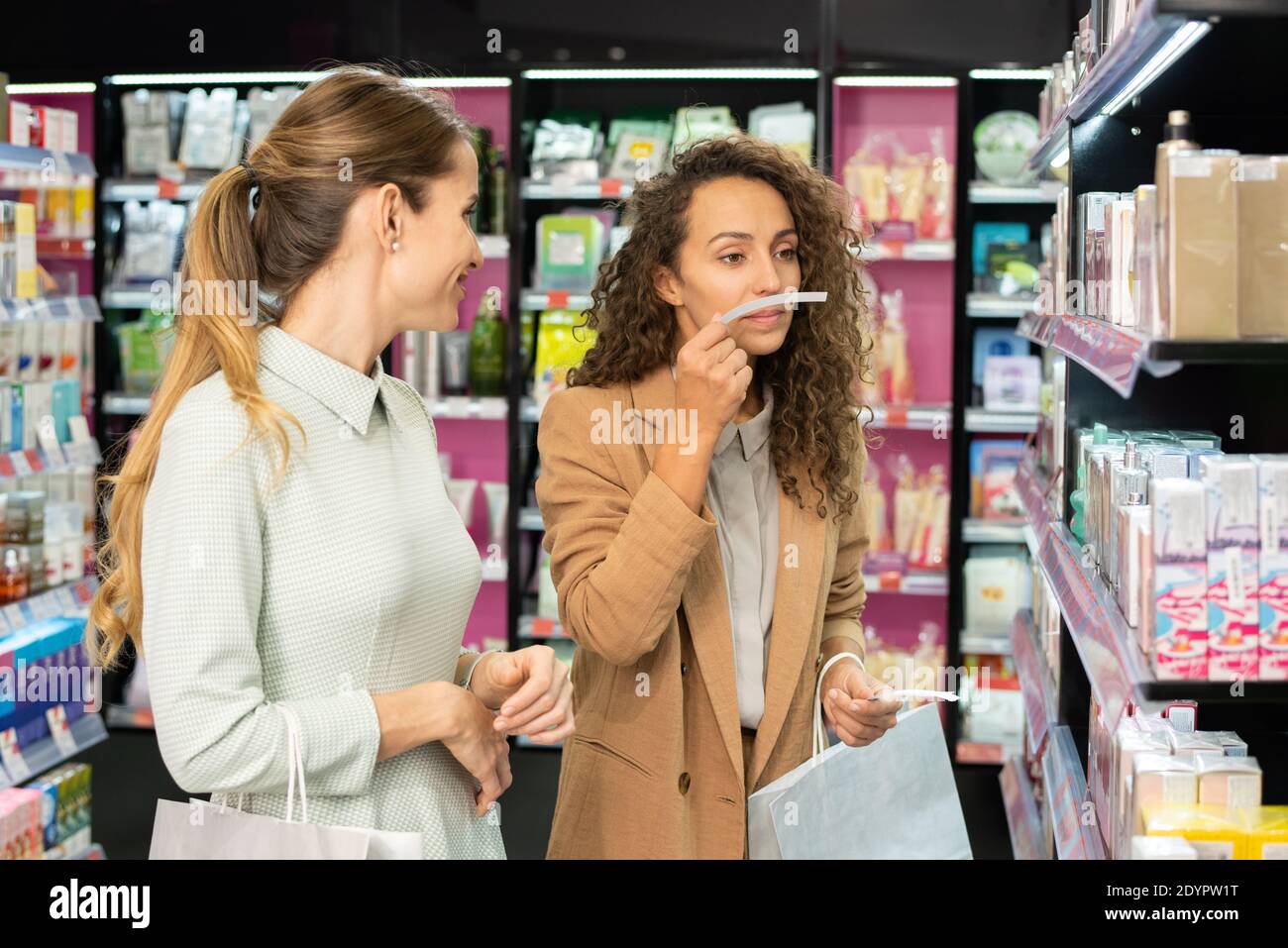Young woman with curly hair holding paper blotter by her nose while smelling new fragrance with her friend in beauty department Stock Photo