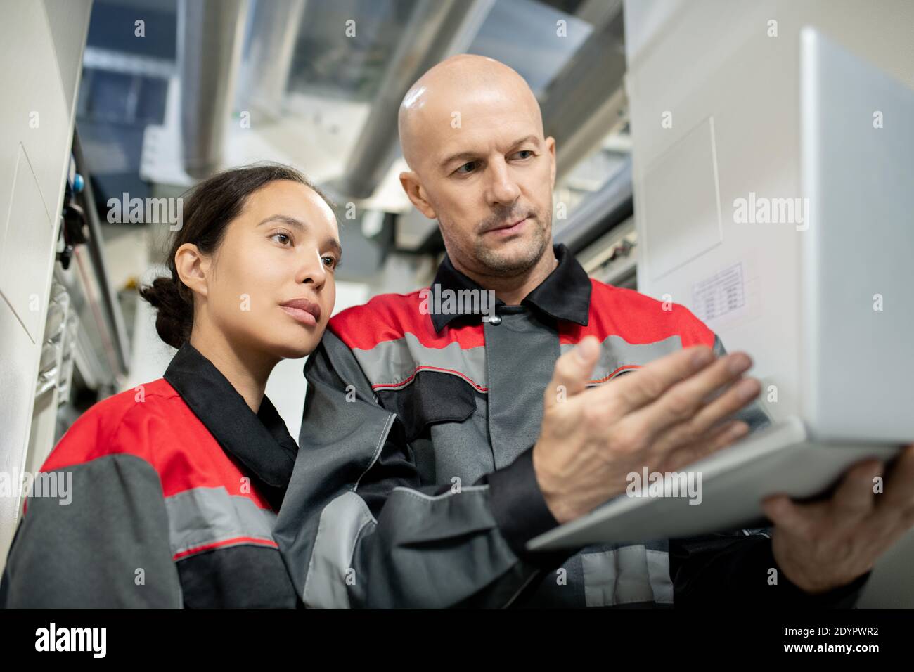 Serious bald mature foreman in workwear holding laptop and pointing at data on display while making presentation to his young female subordinate Stock Photo