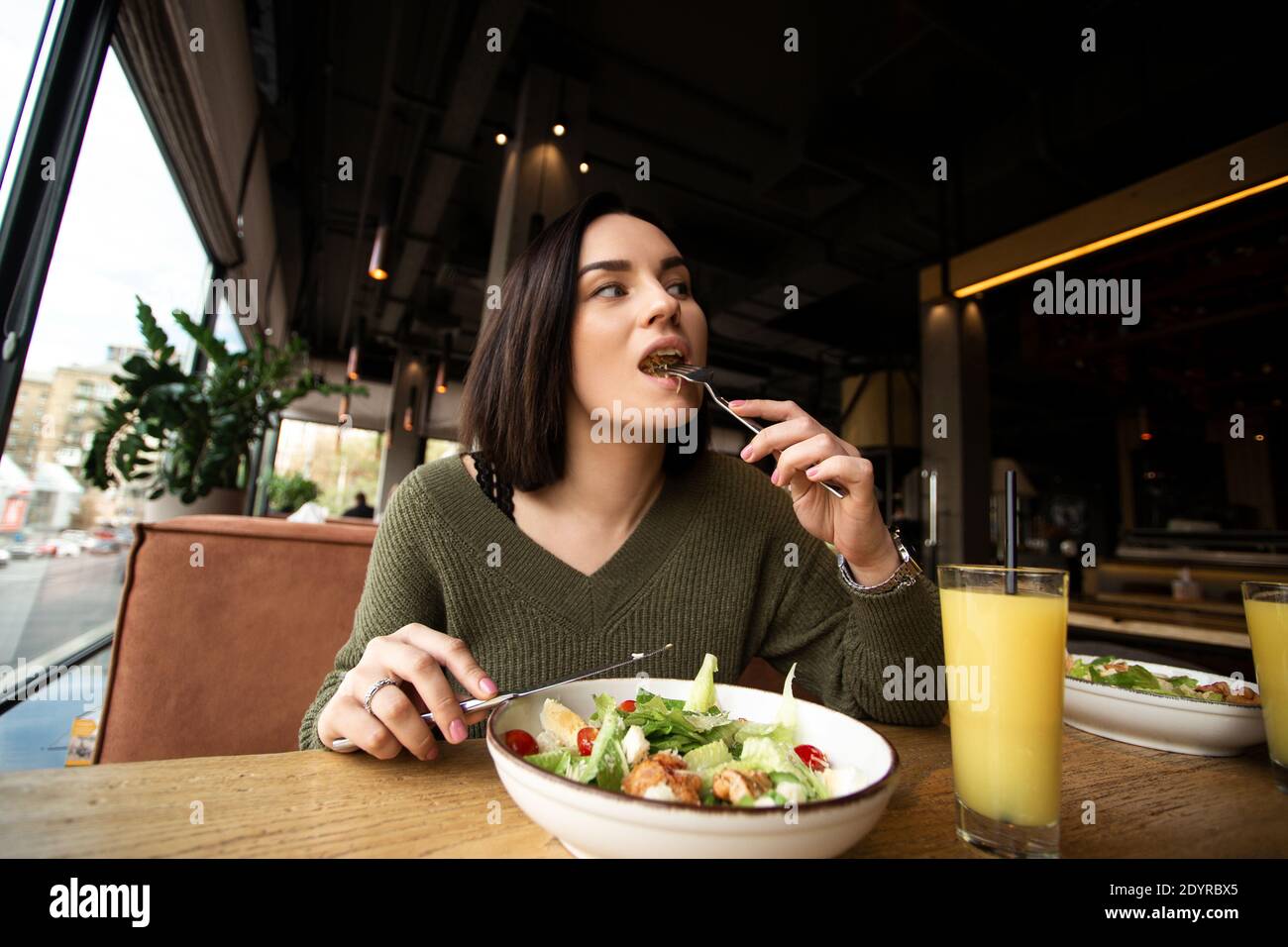 Young woman enjoys tasty meal. Attractive woman with brown hair slowly eating healthy caesar salad and looking away. Glass of orange juice. Restaurant Stock Photo
