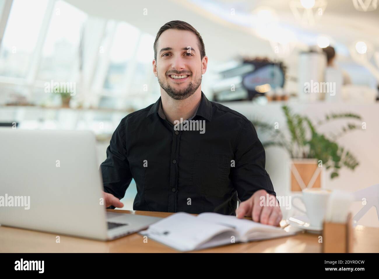 Cheerful freelancer man working from cafe. Attractive young man smiling, using his modern laprtop and looking at camera. Cozy coffeehouse on Stock Photo