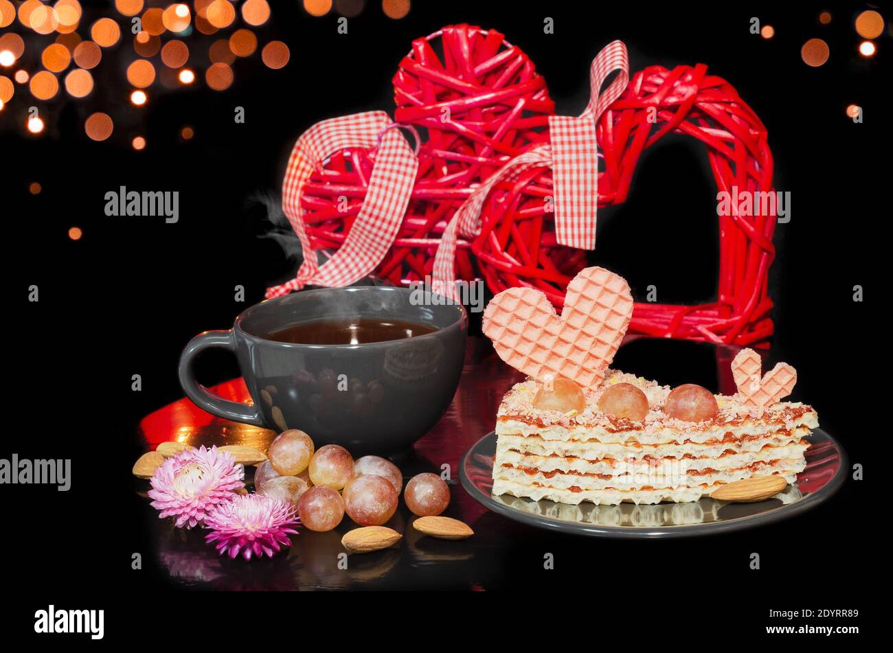 Waffle cake decorated with a heart and grapes and hot tea in a Cup. Black background with bokeh and hearts. Valentine's day concept, selective focus Stock Photo