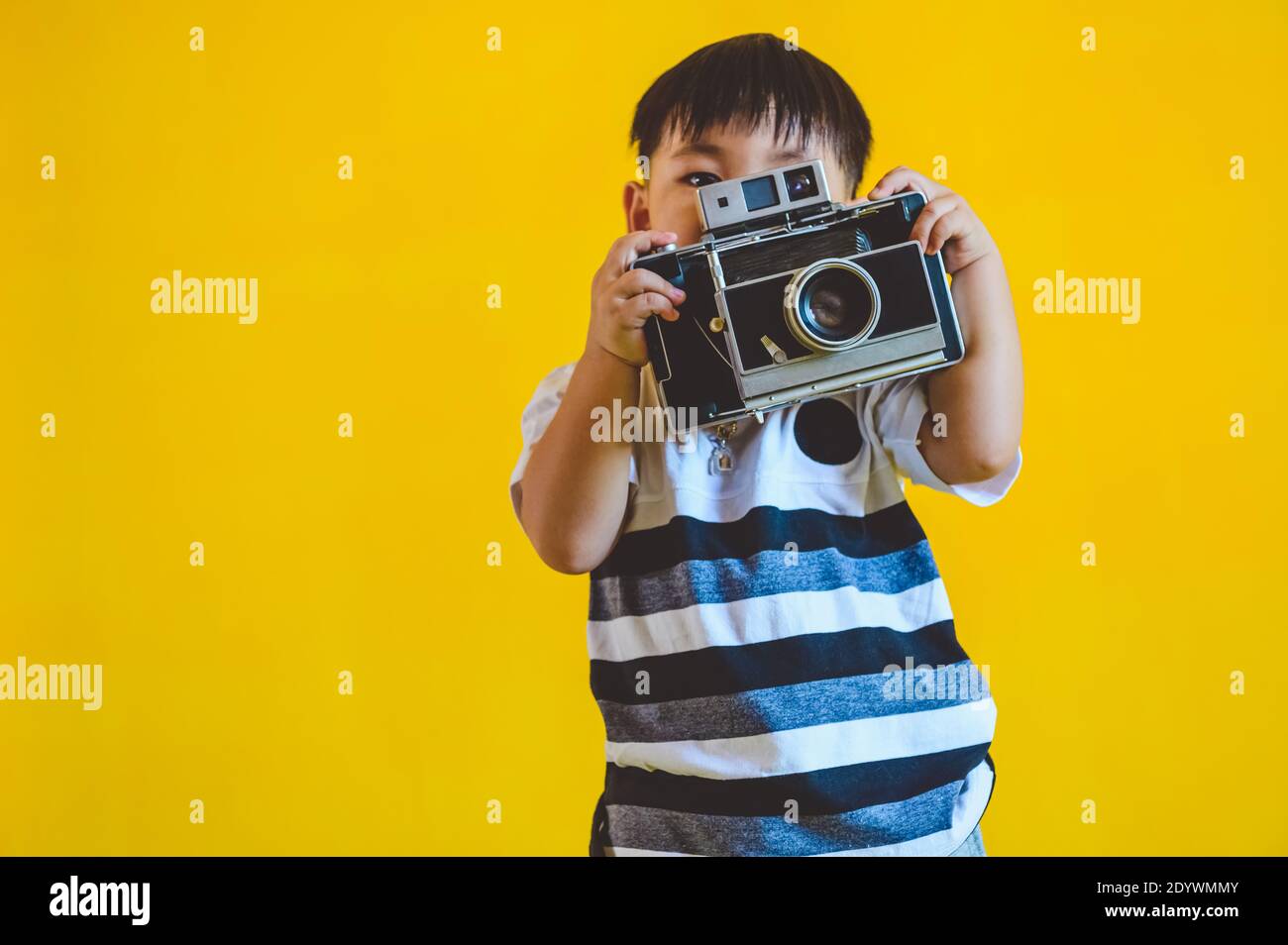 Asian boy holding old vintage camera and posing as photography on yellow isolated background. People lifestyle and technology concept. Stock Photo