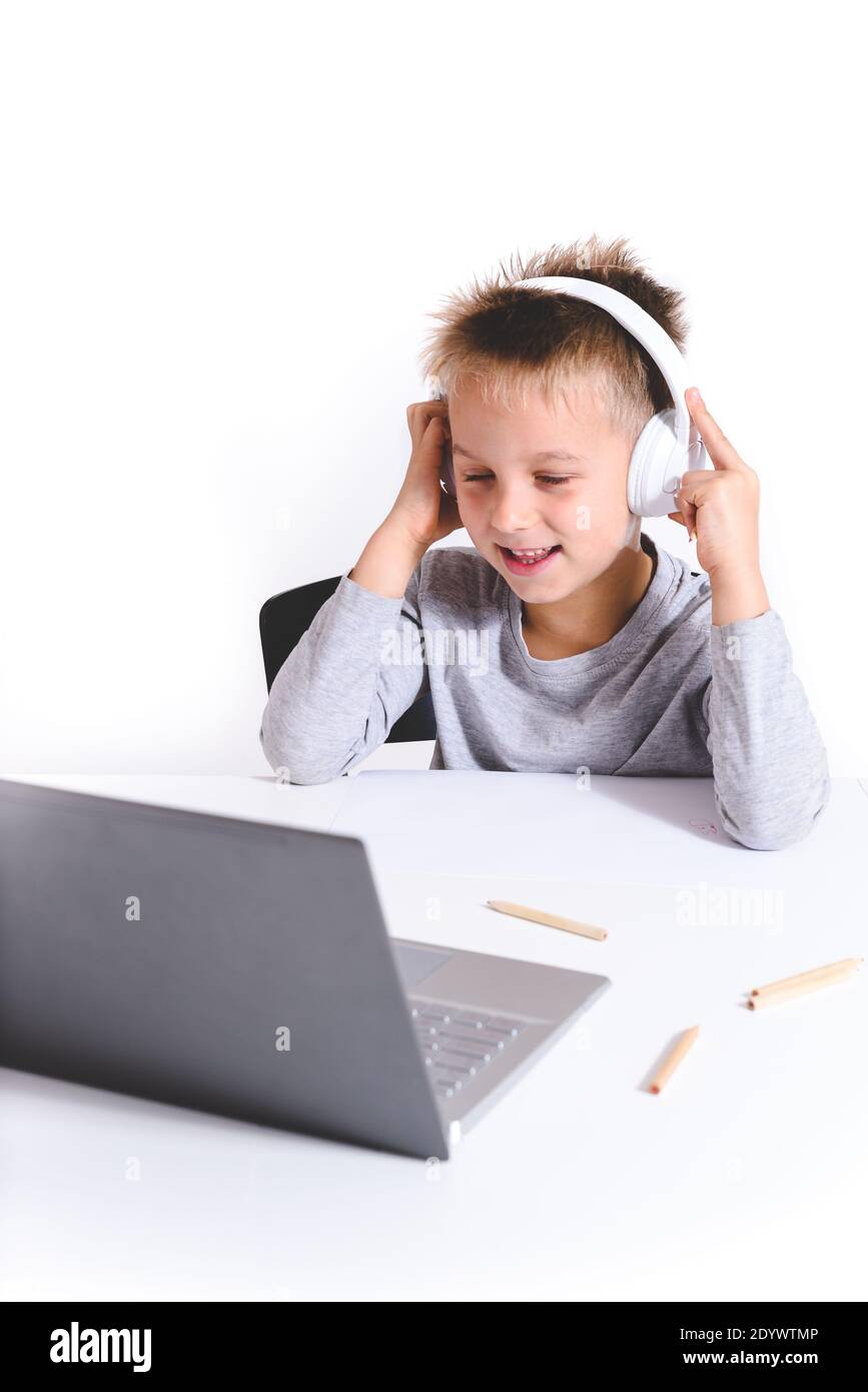 vertical portrait of a blond caucasian kid with headphones looking to the screen of a laptop with white background and table Stock Photo