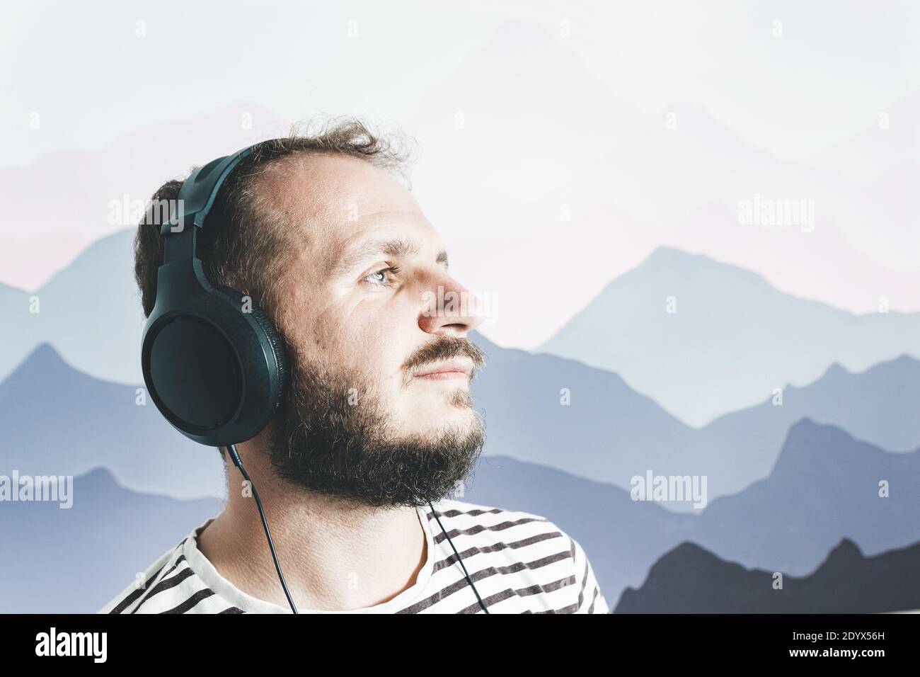 Bearded happy man in a striped T-shirt listens to music with big headphones. looks away. Stock Photo