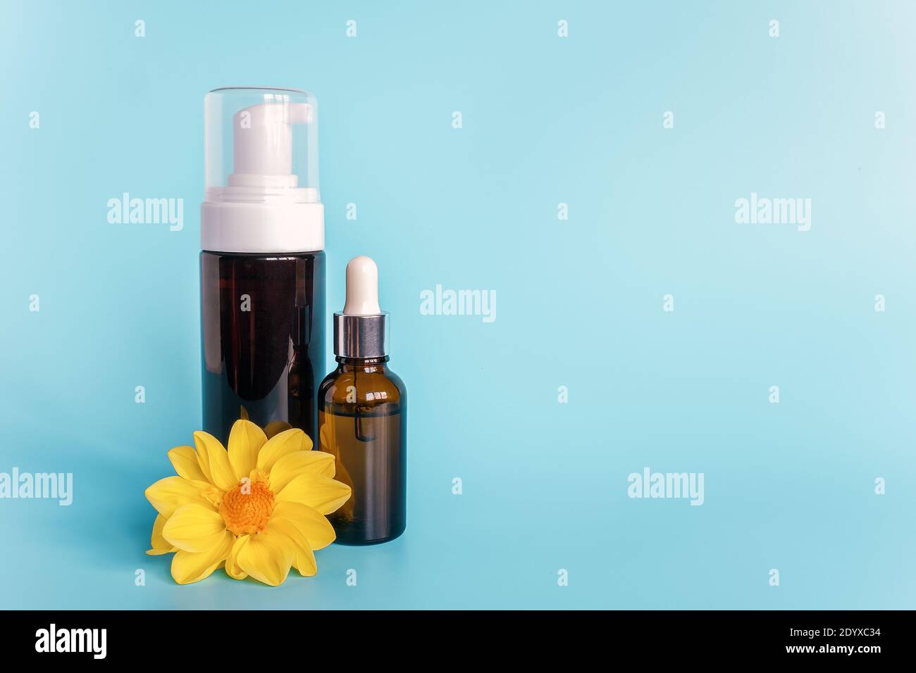 Essential oil in small open brown dropper bottle with lying glass pipette, big bottle with white dispenser and yellow flower on blue background. Conce Stock Photo