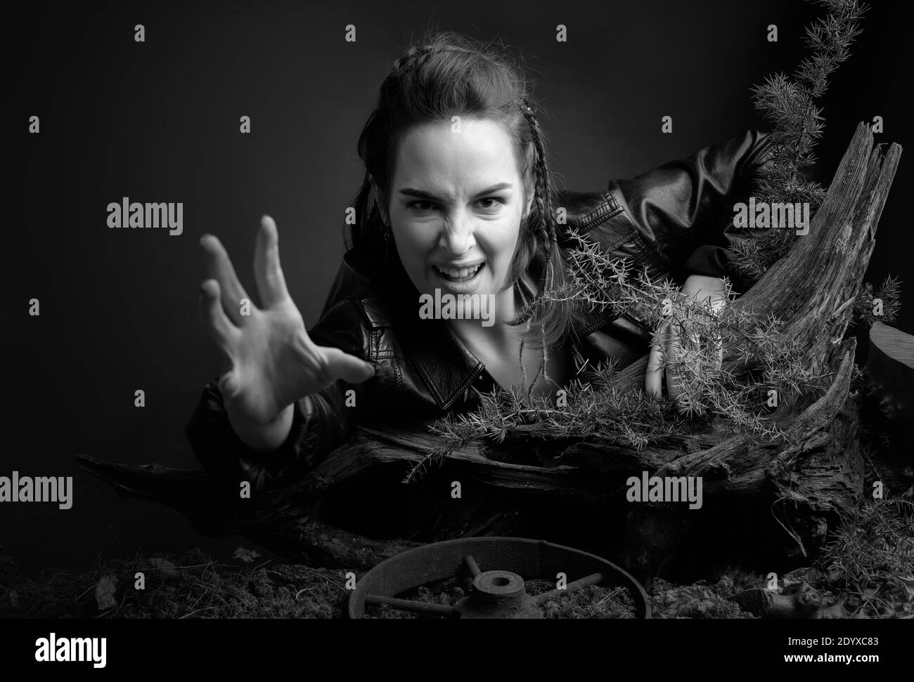 A young woman in a black leather jacket climbs out of the pit holding on to a snag. Angry girl stretches out her hand to the camera. Expressive facial Stock Photo