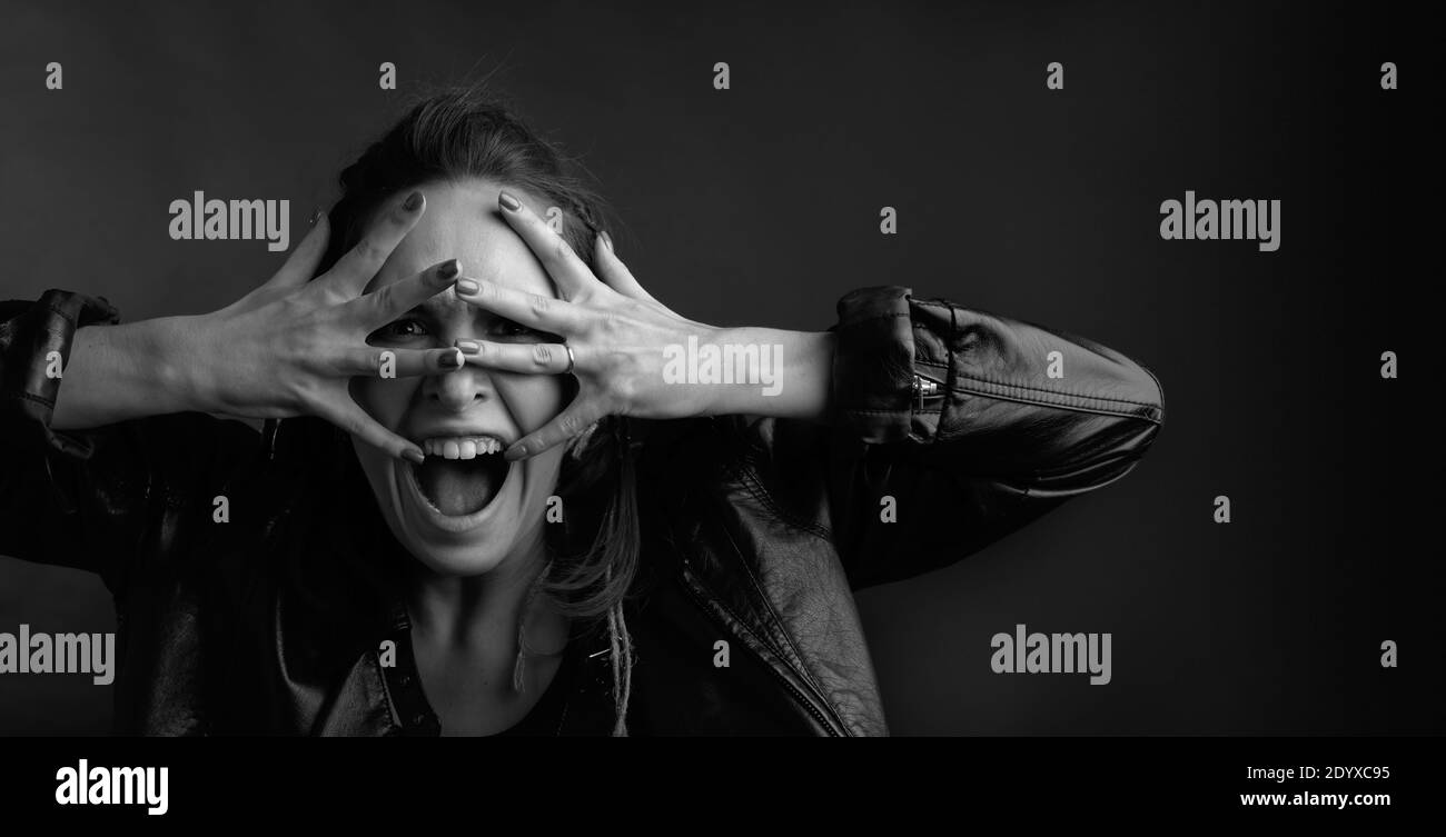Portrait of a displeased woman wearing a black leather jacket. Screaming woman covers her face with her hands. Black and white. Stock Photo