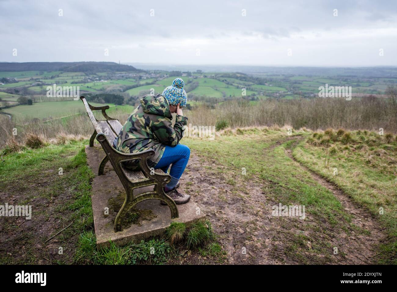 Sad woman sitting alone on a bench in the countryside looking across fields and hills on a lockdown walk in the winter Stock Photo