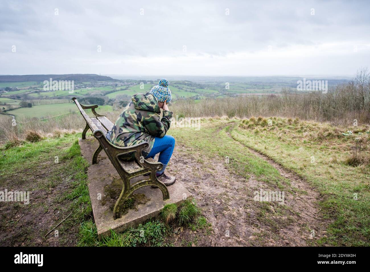 Sad woman sitting alone on a bench in the countryside looking across fields and hills on a lockdown walk in the winter Stock Photo