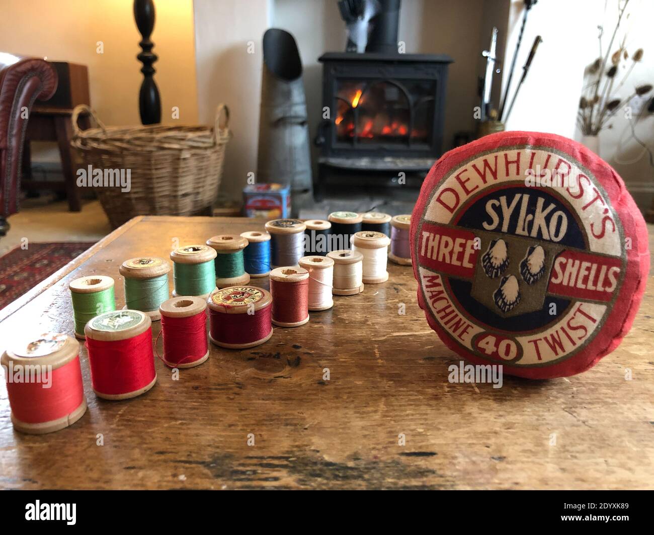 Colourful reels of cotton and a vintage Dewhurst Sylko pincushion lined up on a box infront of a cosy living room with a woodburner Stock Photo