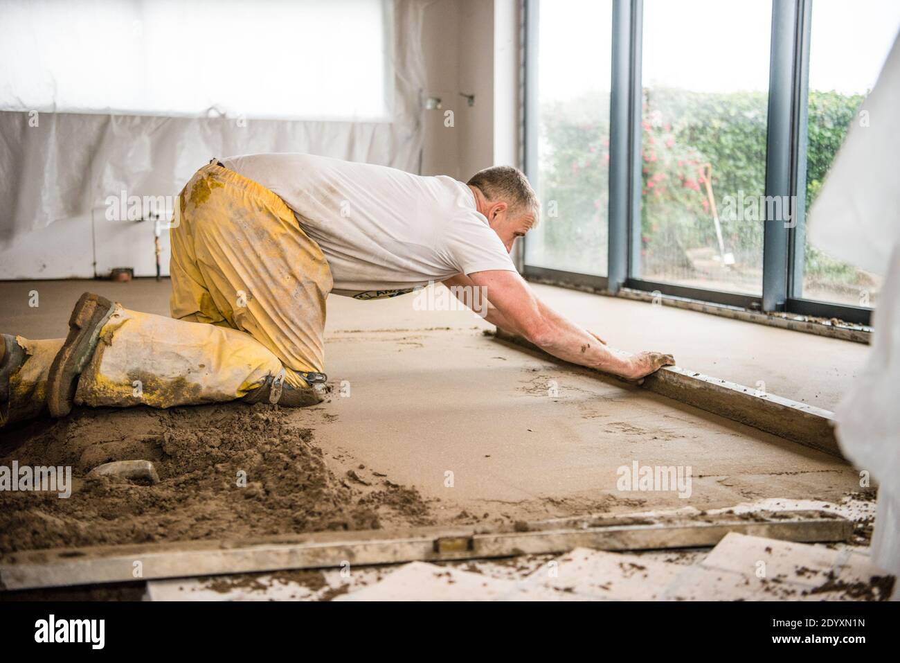 White middle-aged tradesman in yellow overalls concentrating screeding a new floor in a house extension for a new kitchen Stock Photo