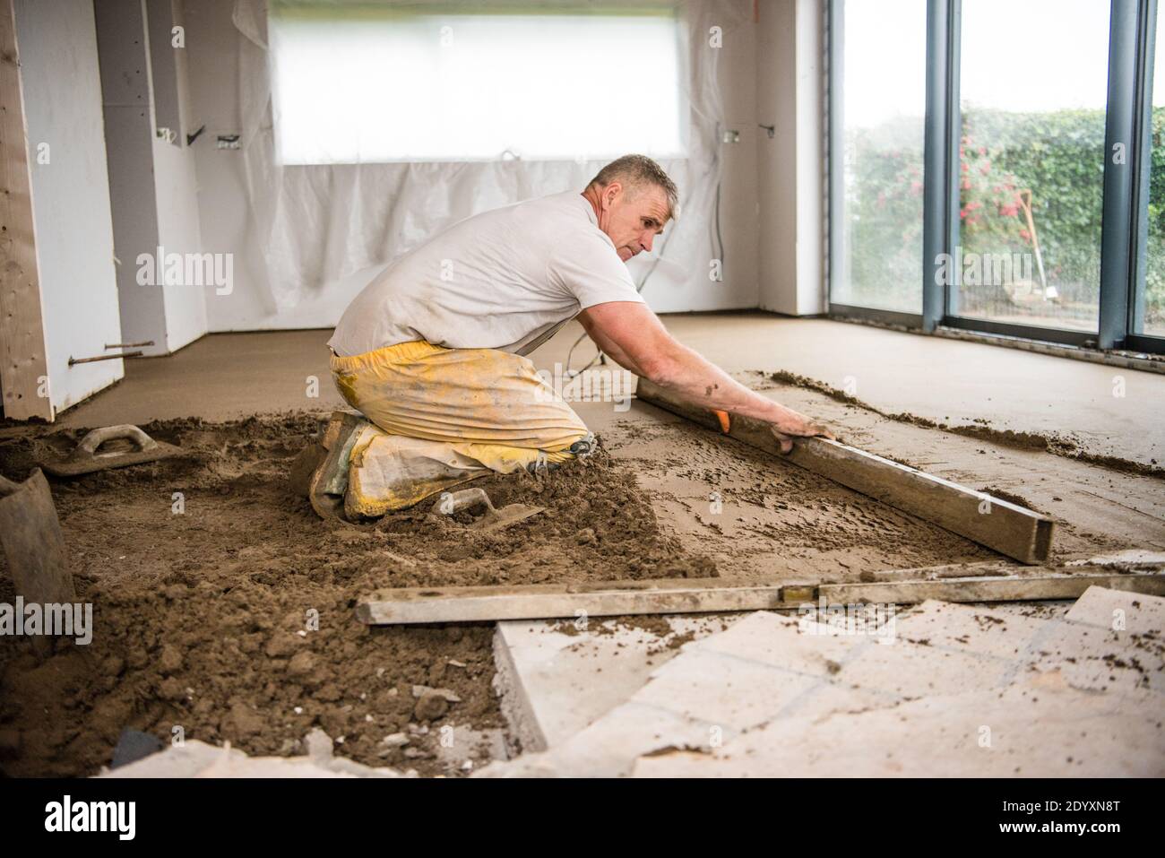 White middle-aged tradesman in yellow overalls concentrating screeding a new floor in a house extension for a new kitchen Stock Photo