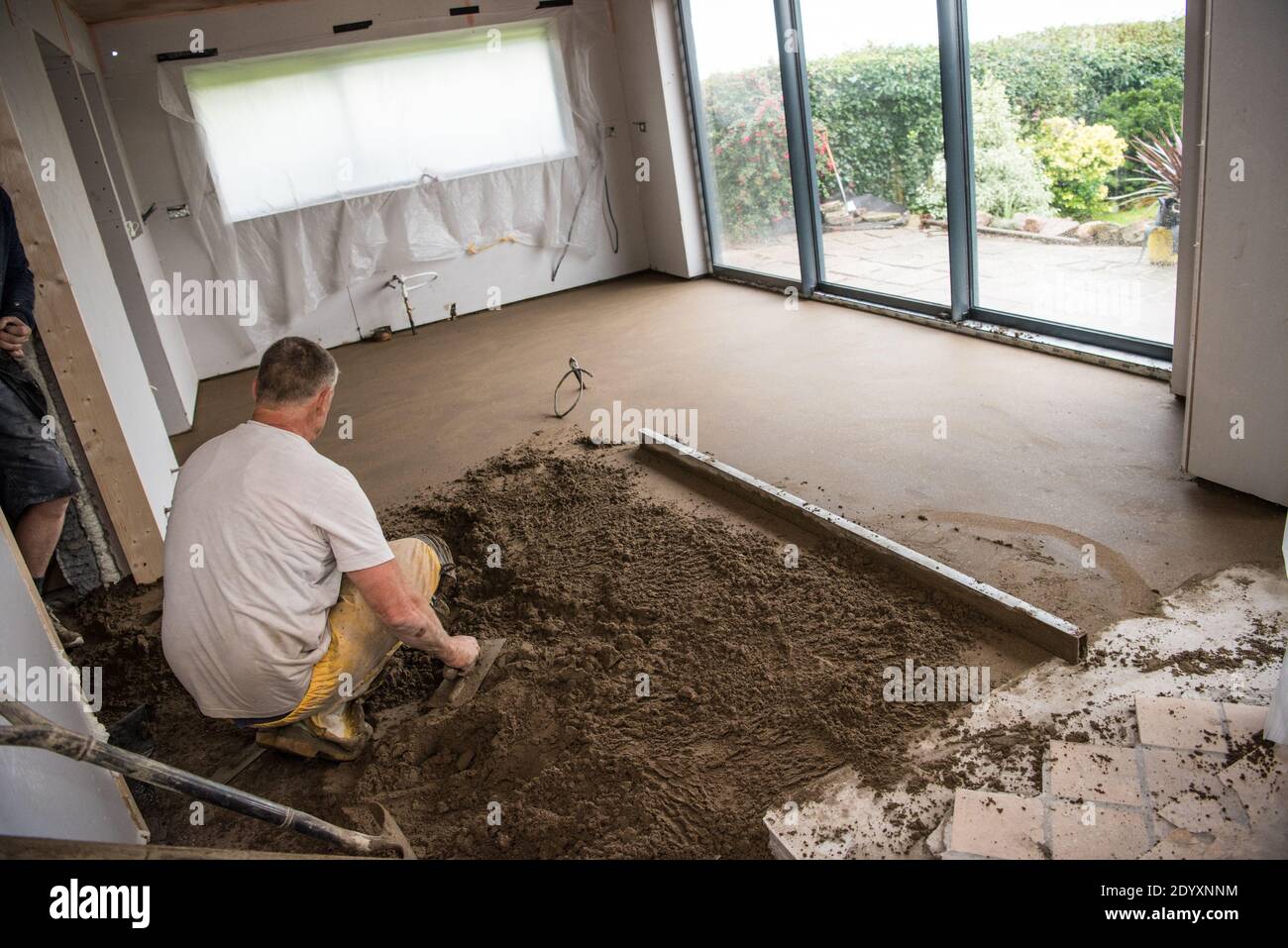 White middle-aged tradesman in yellow overalls concentrating screeding a new floor in a house extension for a new kitchen Stock Photo