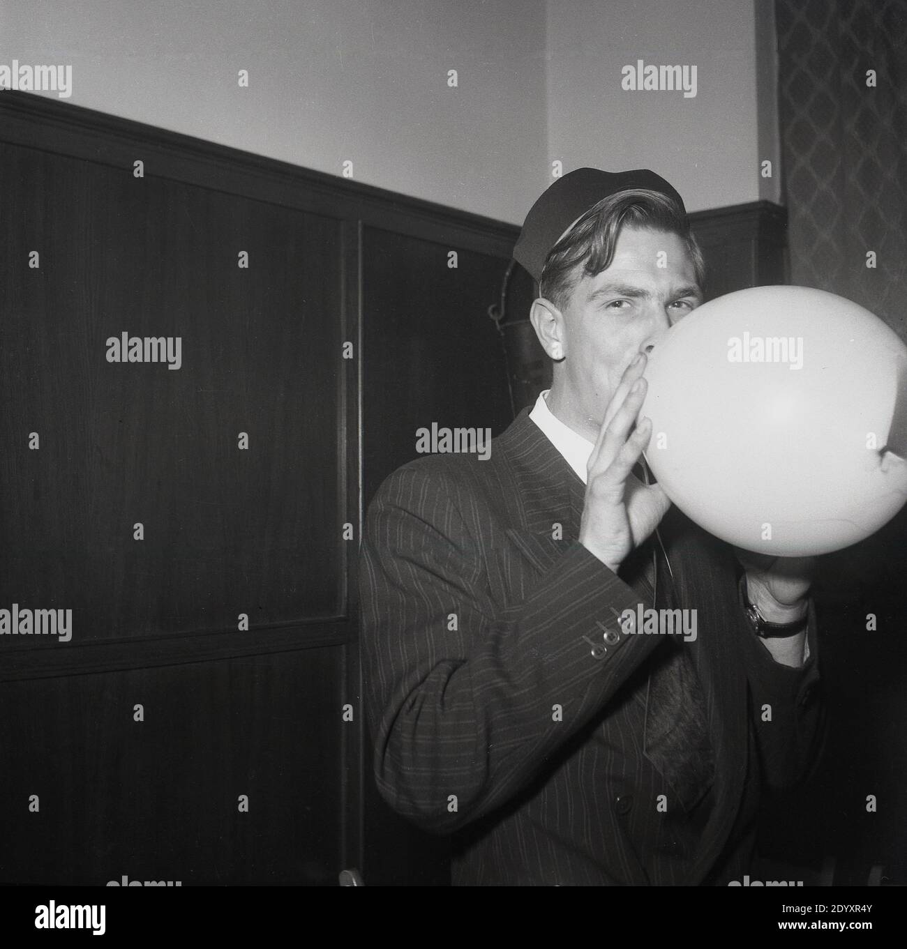 1950s, historical, party time....a man in a pinstripe suit and wearing a party hat in a function room blowing up a ballon, England, UK. Stock Photo