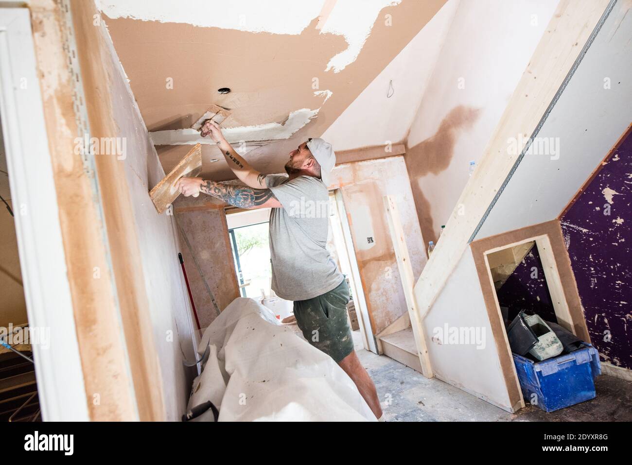 White tattooed man wearing a baseball cap plastering walls and ceiling on a house renovation project Stock Photo