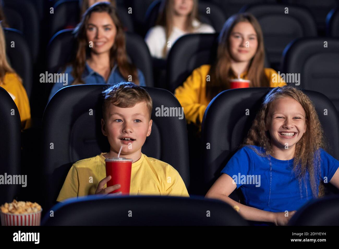 Front view of caucasian kids enjoying time together holding sparkling drink, young audience on background. Happy little friends sitting in cinema, watching funny cartoon. Entertainment concept. Stock Photo