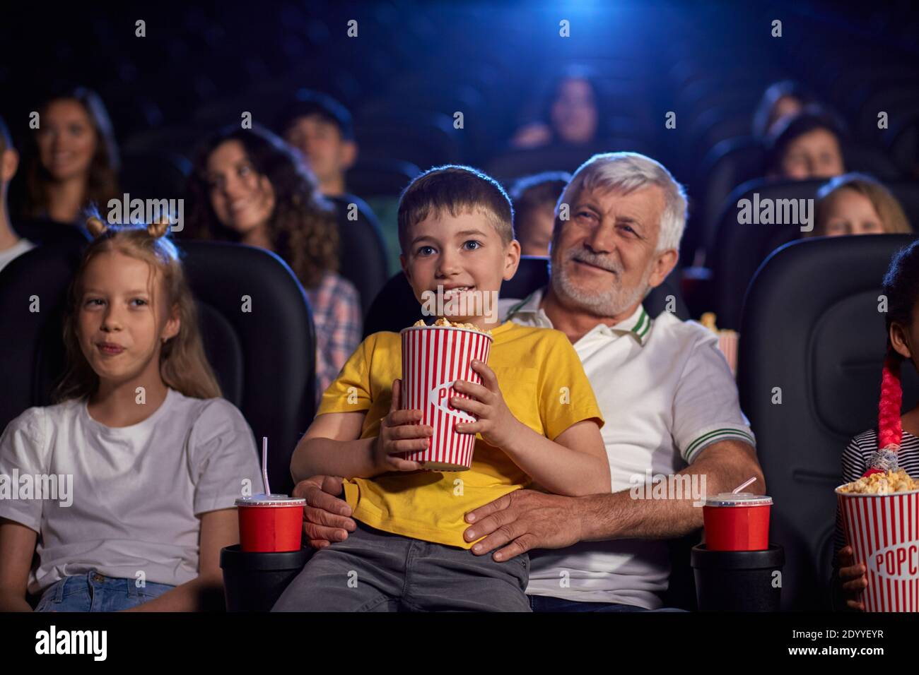 Selective focus of bearded senior grandfather enjoying time with grandson, kid sitting on knees and holding popcorn bucket. Happy caucasian family sitting in cinema, watching funny cartoon. Stock Photo