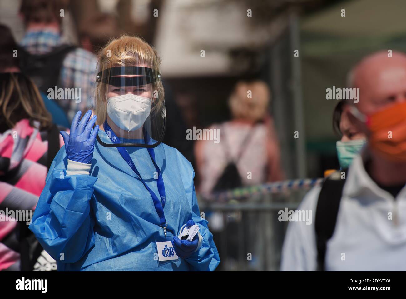 Medic wearing a respirator and plastic face shield waving her hand next to a queue of people waiting for a corona virus test. Stock Photo