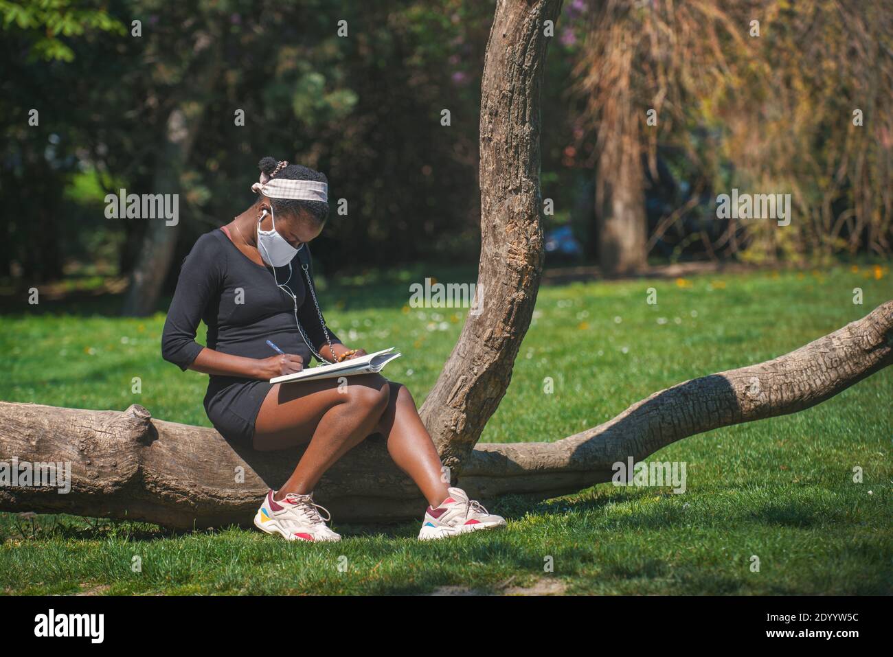 Woman with a cloth face mask sitting on a lying tree trunk writing in a questionnaire before passing a test against the corona virus. Stock Photo