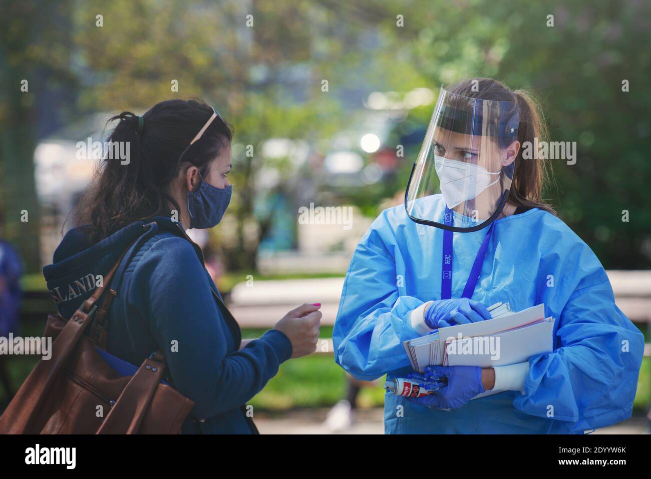Medic with respirator and plastic face shield giving a questionnaire to a woman with cloth face mask before undergoing a corona virus test. Stock Photo