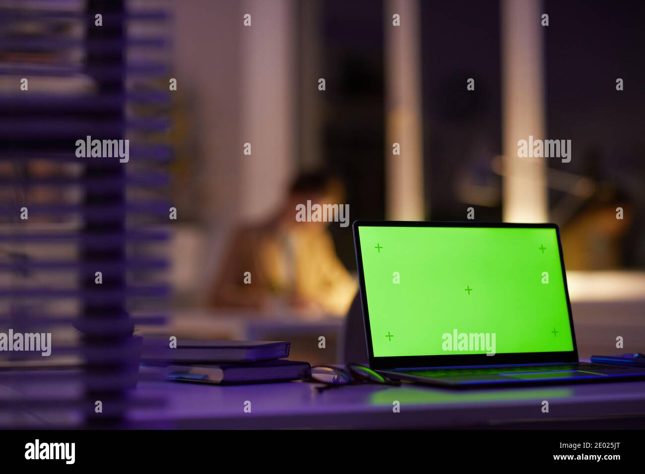 Modern office workplace late in evening with green screen laptop on foreground and unrecognizable woman working on background, copy space Stock Photo