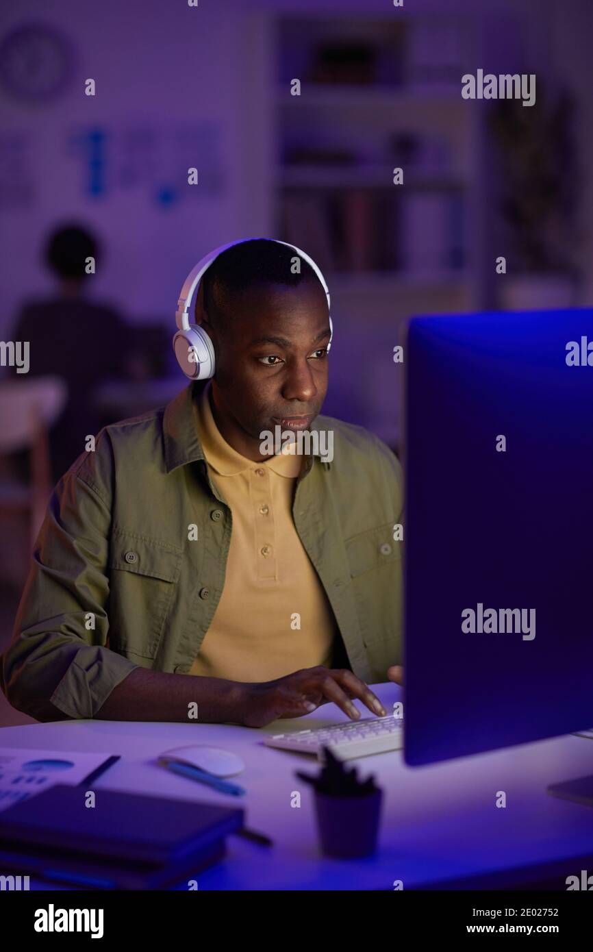 Portrait of African American man wearing casual outfit listening to music in headphones while working on computer in evening Stock Photo