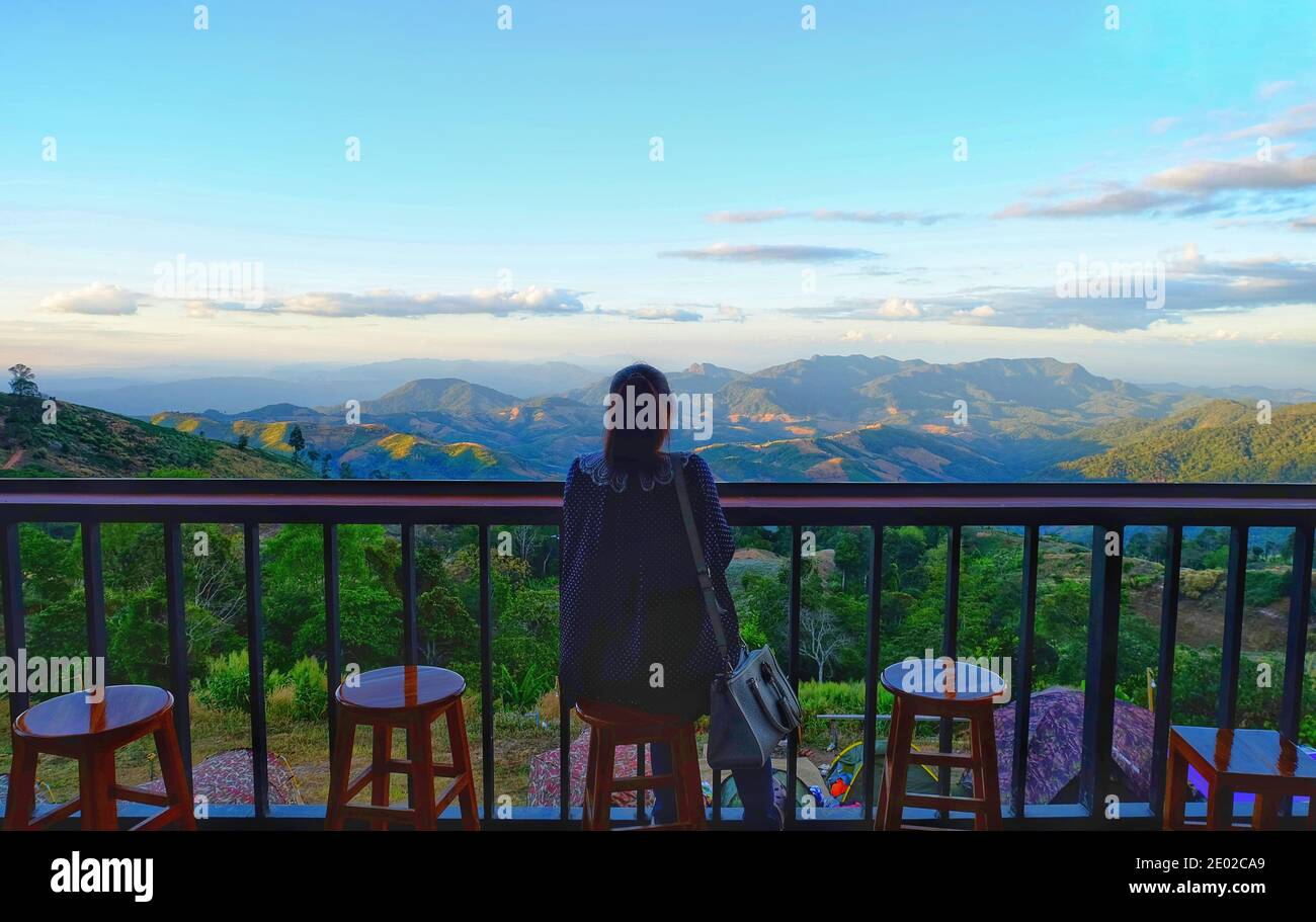 A back view of an Asian woman sitting on a wooden stool on a ledge at a roadside view point of a green tropical mountain range in northern Thailand Stock Photo