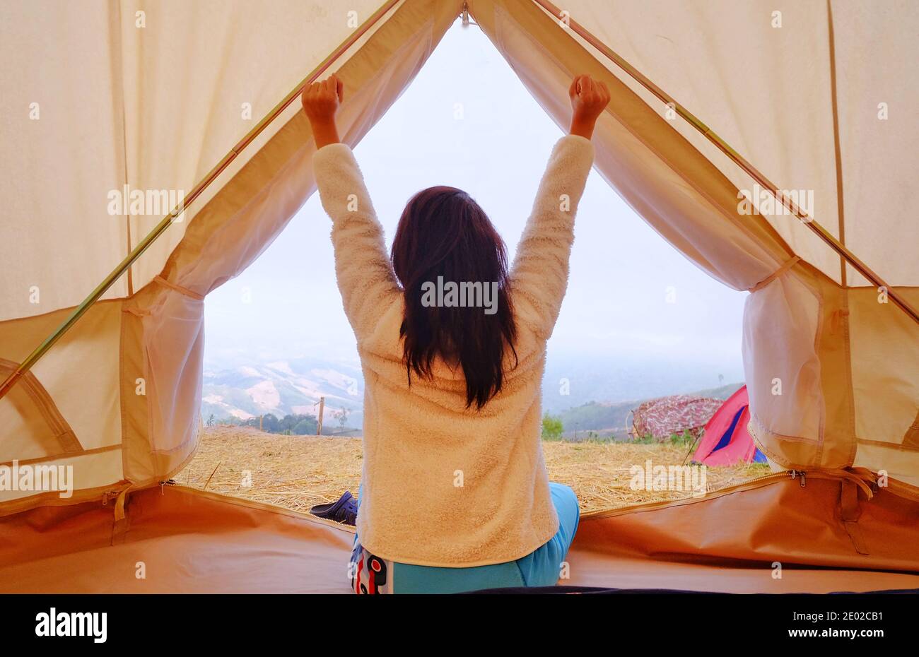 A young woman sitting peacefully at inside her white tent in the morning with cool breeze and morning sun, stretching and relaxing. Stock Photo