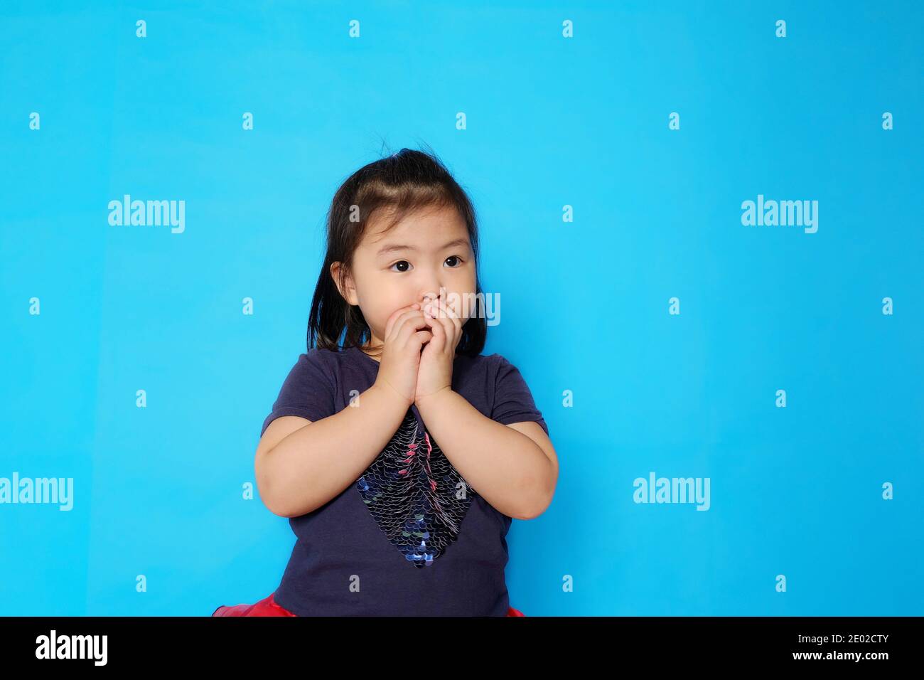 A cute Asian girl covering her mouth with her hands with surprise. Plain light blue background. Stock Photo