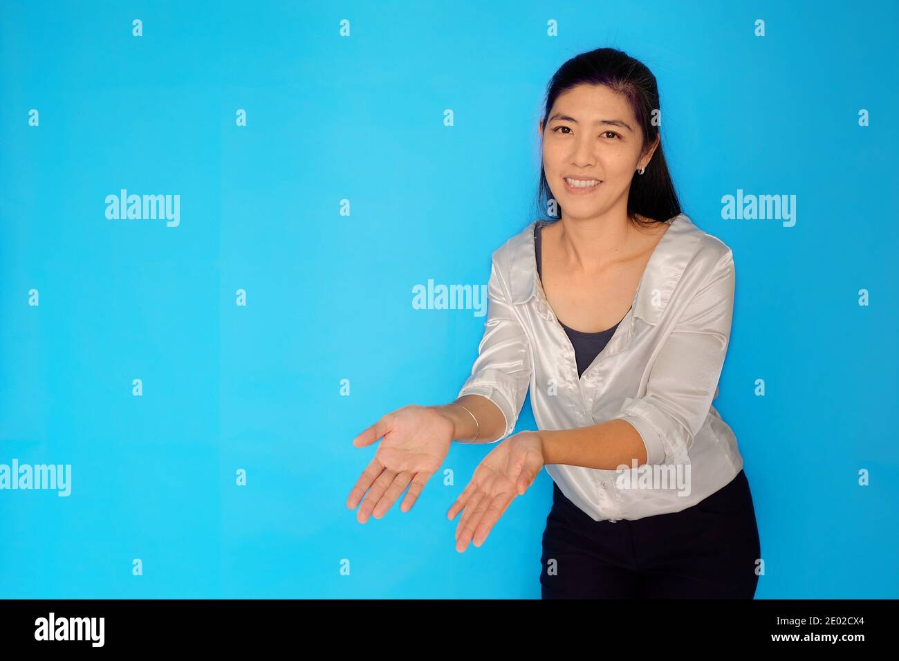 A waist height shot of an attractive Asian woman in her 30s presenting a product, pointing with open hands on a light blue plain background. Stock Photo