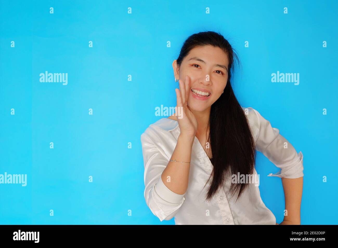 An attractive Asian woman telling a secret by whispering the news quietly, playfully holding her hand close to her mouth, smiling and happy with plain Stock Photo