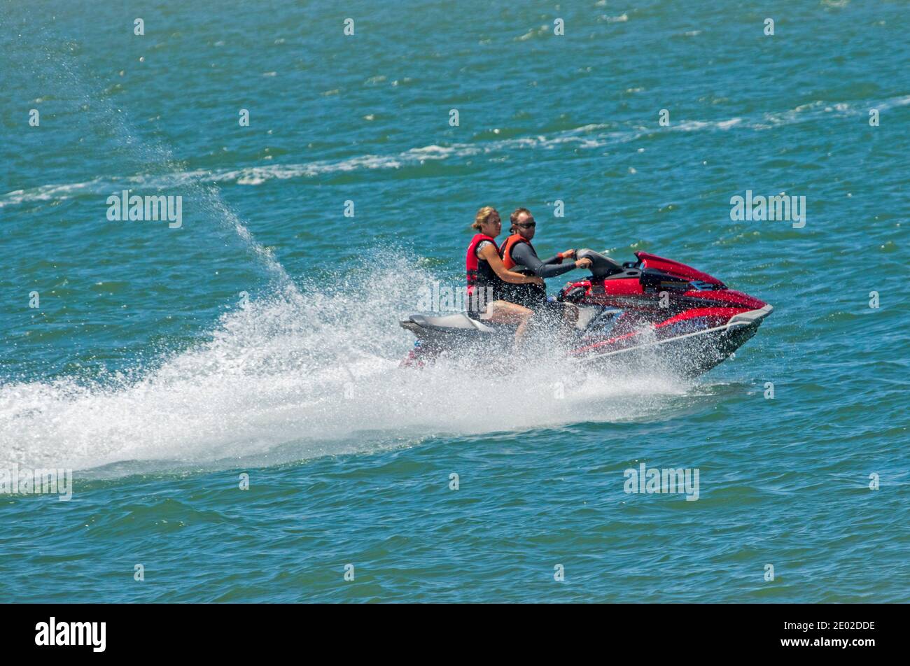 Two people riding bright red jet ski on blue water of Pacific Ocean with water spraying into air, in Queensland Australia Stock Photo