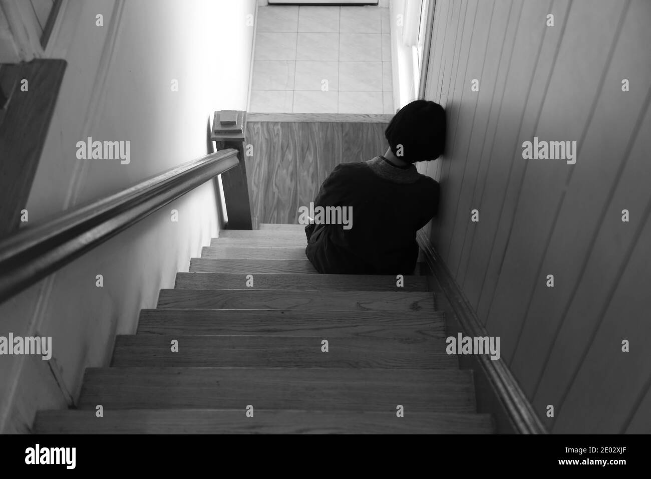 Woman sitting alone sadly on wooden staircase, leaning her head against the wall, back view, white tone Stock Photo