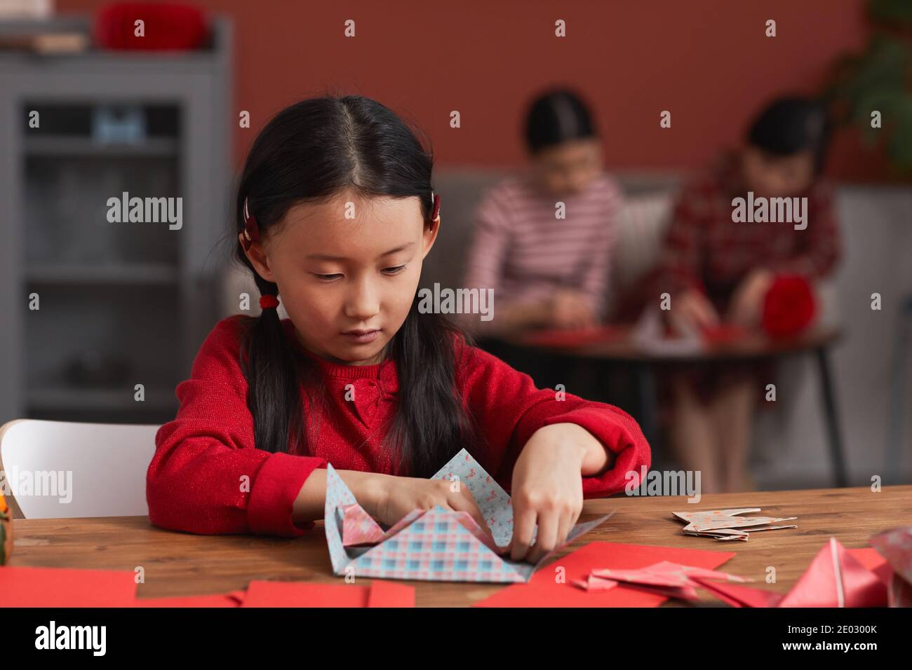 Cute little Asian girl wearing red clothes sitting at wooden table in living room busy making paper craft for Lunar New Year decorations Stock Photo