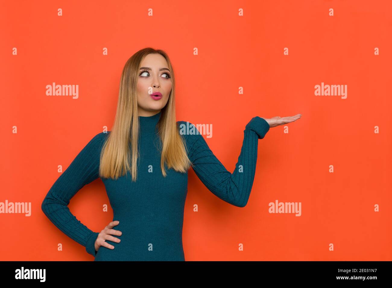 Surprised young woman in teal sweater is holding hand raised and looking at the side. Waist up studio shot on orange background. Stock Photo