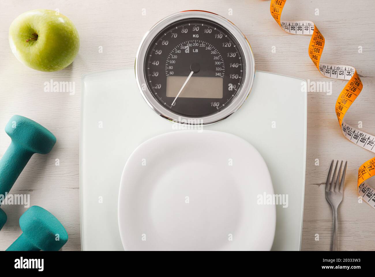 Weight control concept with plate on scale and cutlery apple and dumbbells on white wooden table. Top view. Horizontal composition. Stock Photo