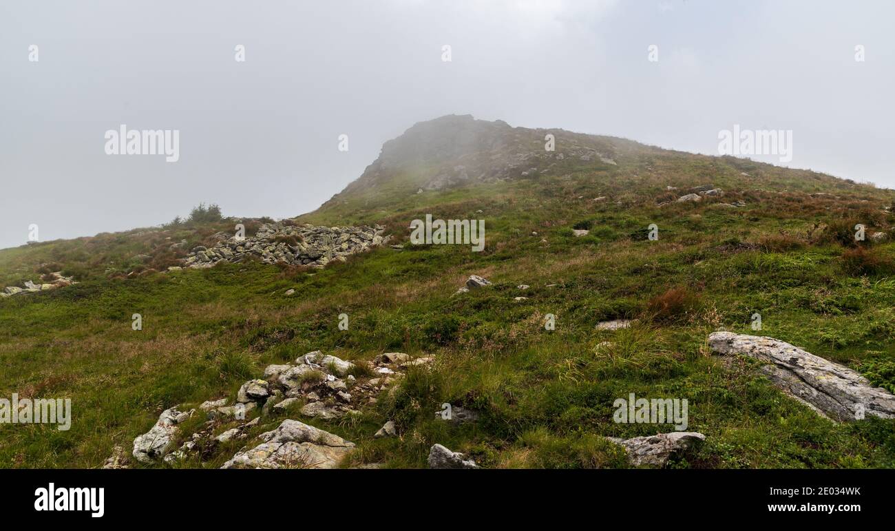 Hill covered by meadow with stones and rock formation on summit during cloudy day - Varful Mutu hill in Valcan mountains above Straja ski resort in Ro Stock Photo