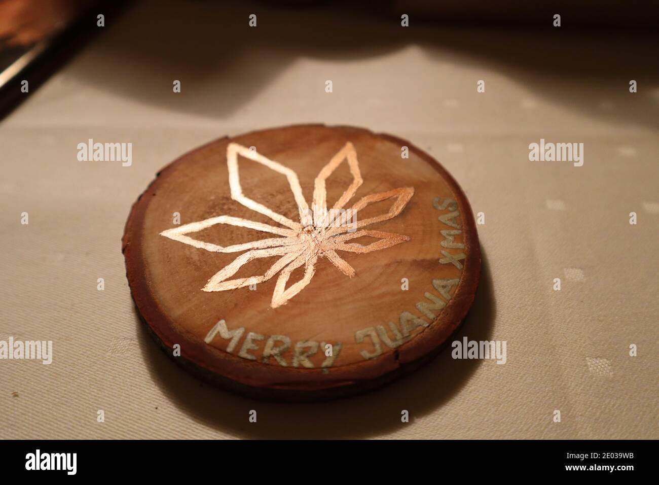 Selfmade wooden coaster on a table with a marijuana leaf in bright colors. The text MERRY JUANA XMAS decorates the coaster. Stock Photo