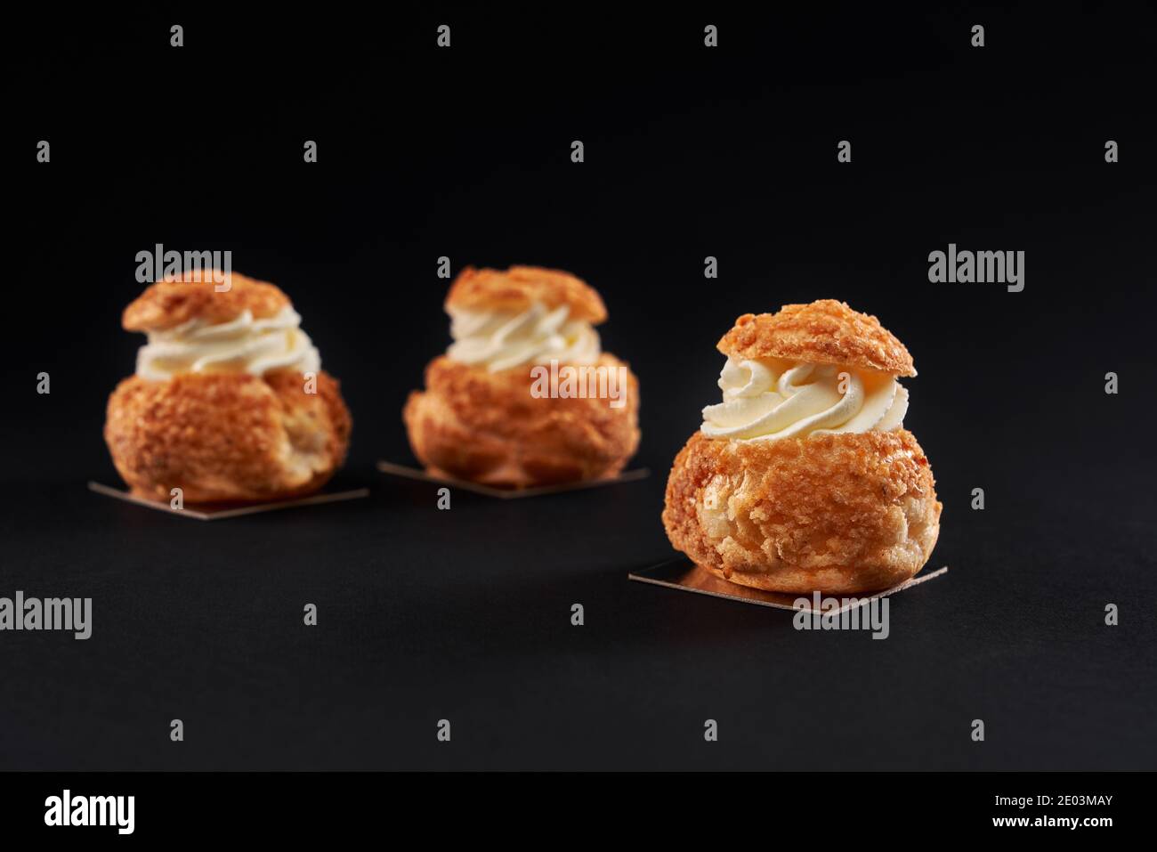 Three delicious fresh crunchy profiteroles with sweet white cream inside. Closeup of homemade tasty eclairs isolated on black background. Concept of desserts, restaurant food. Stock Photo