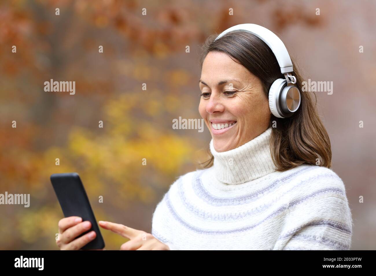 Happy adult woman using headphones and smart phone listening to music in a park in autumn Stock Photo