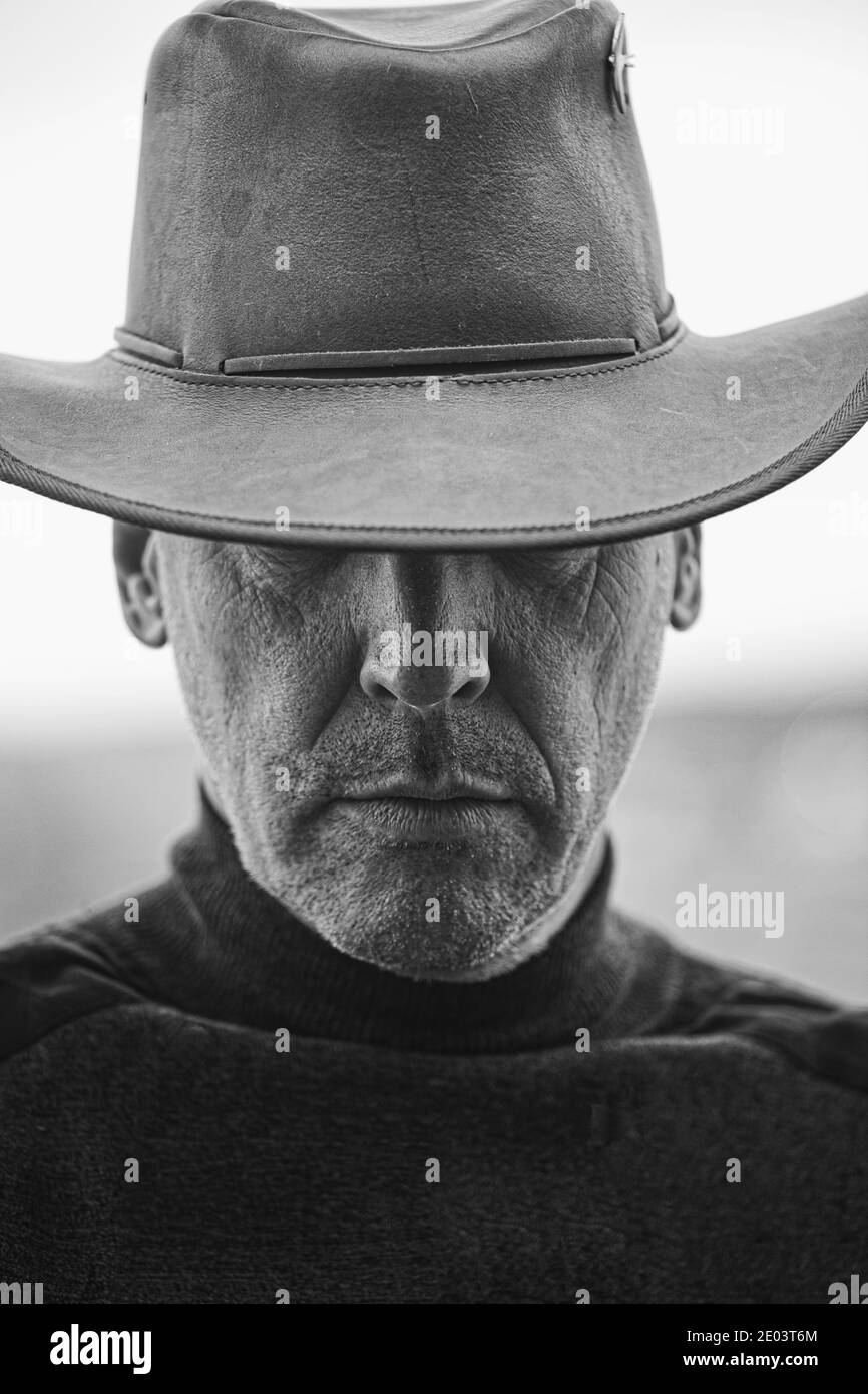 Black and white image of middle aged unshaven man with serious expression wearing cowboy hat covering his eyes. Moody, serious, intriguing close up Stock Photo