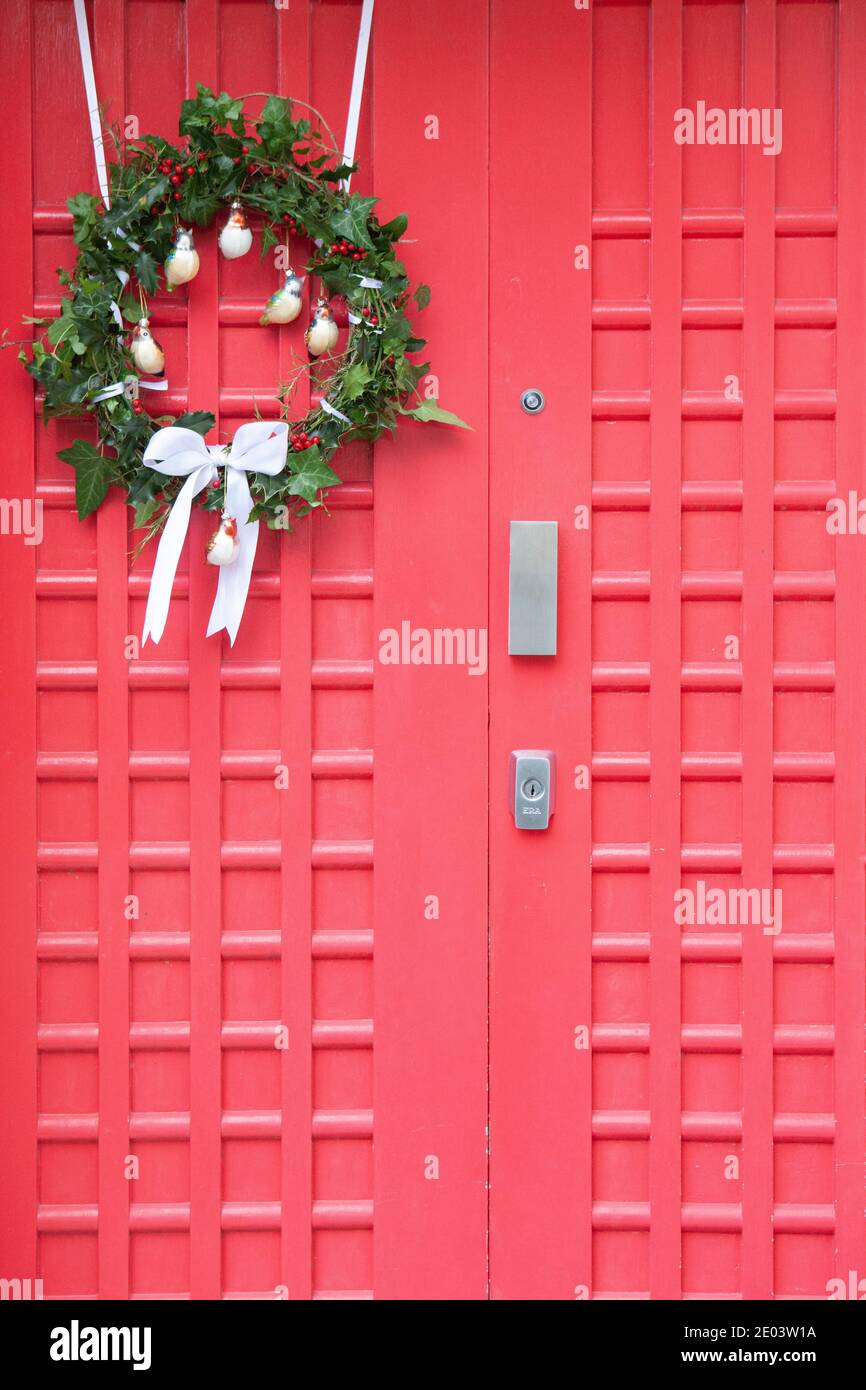 Home-made Christmas wreath on a red door. The wreath uses holly, ivy and rosemary leaves, white ribbon and bird-shaped baubles. Anna Watson/Alamy Stock Photo