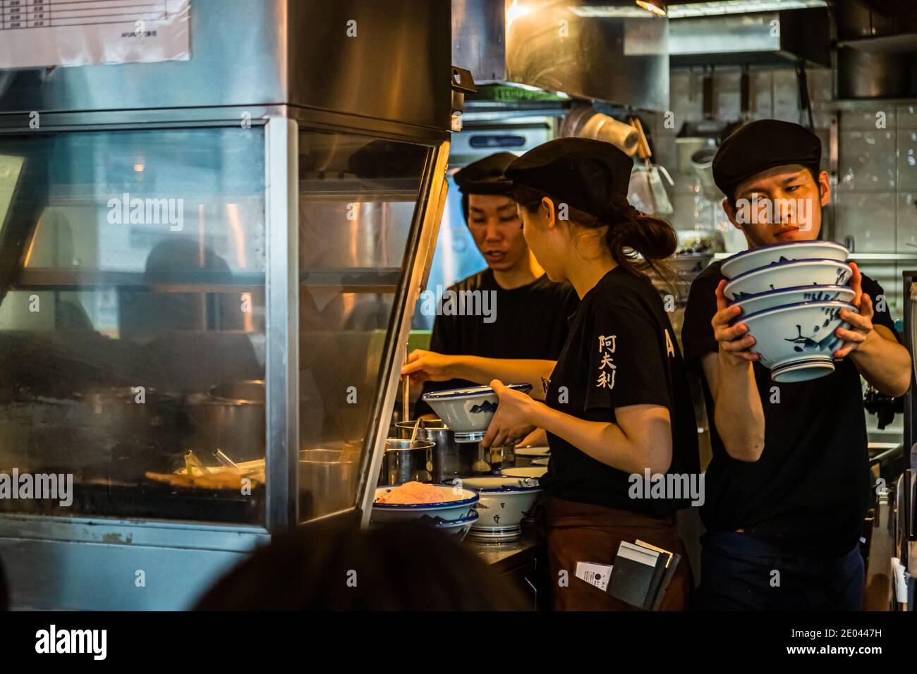 Little space and a lot of activity fill the cramped kitchen of the ramen restaurant Afuri in Shibuya, Tokyo. Three staff members prepare ramen shoulder to shoulder while a colleague holds a stack of blue-and-white bowls ready. Stock Photo