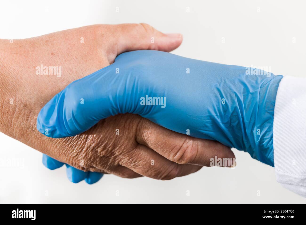 Helping hand of health professional holding old wrinkly palm on white background. Detail of safe handshake of doctor in blue glove and senior patient. Stock Photo